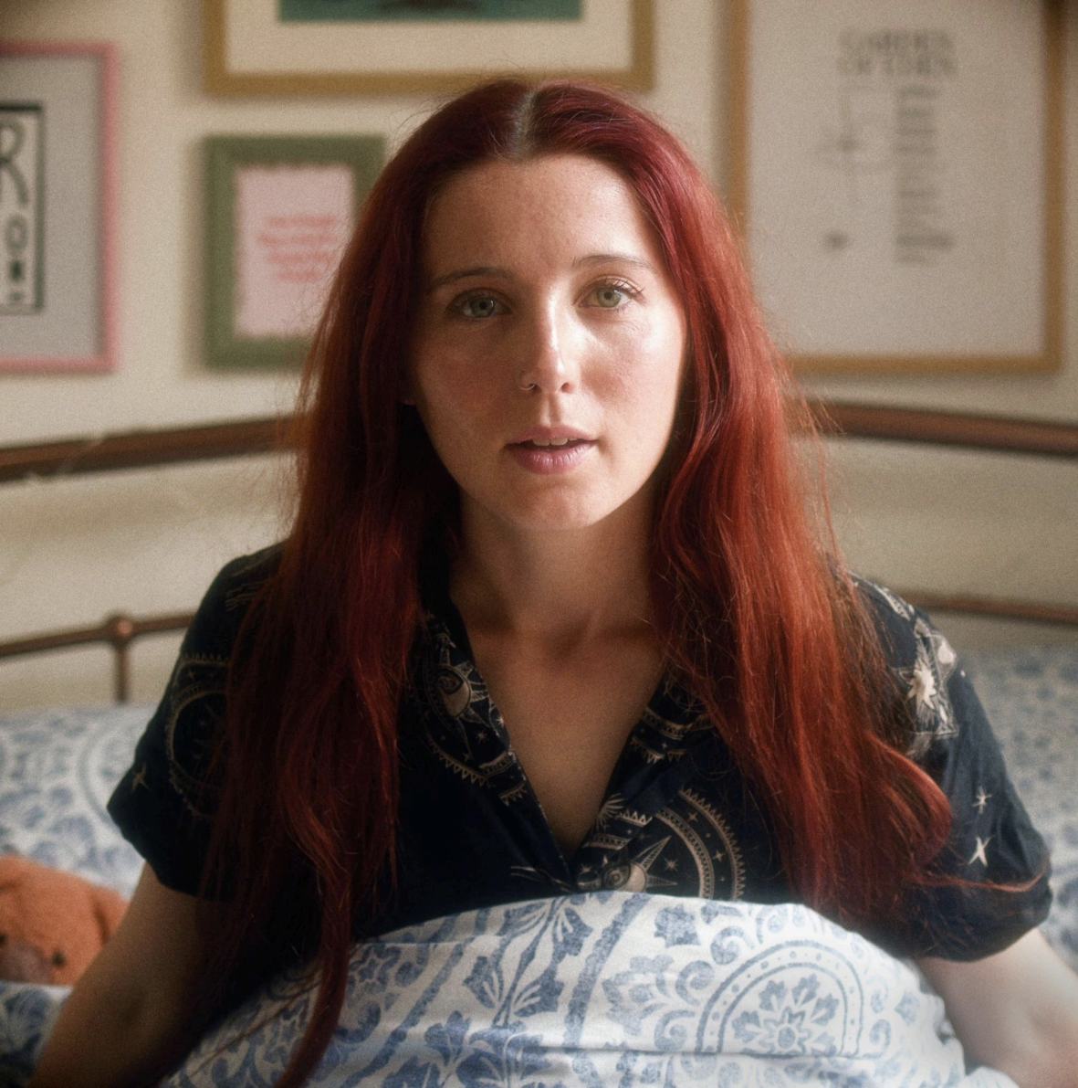alt musician Izzie Derry waking up in her room, with picture frames on the wall behind her.