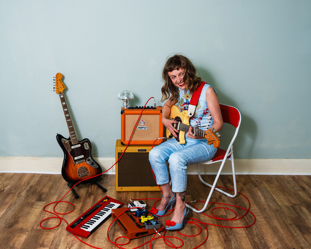 White woman in sleeveless shirt and jeans plays guitar and grins at camera. Another guitar is on a stand, speakers are left of the musician and a keyboard and pedal board are at her feet.