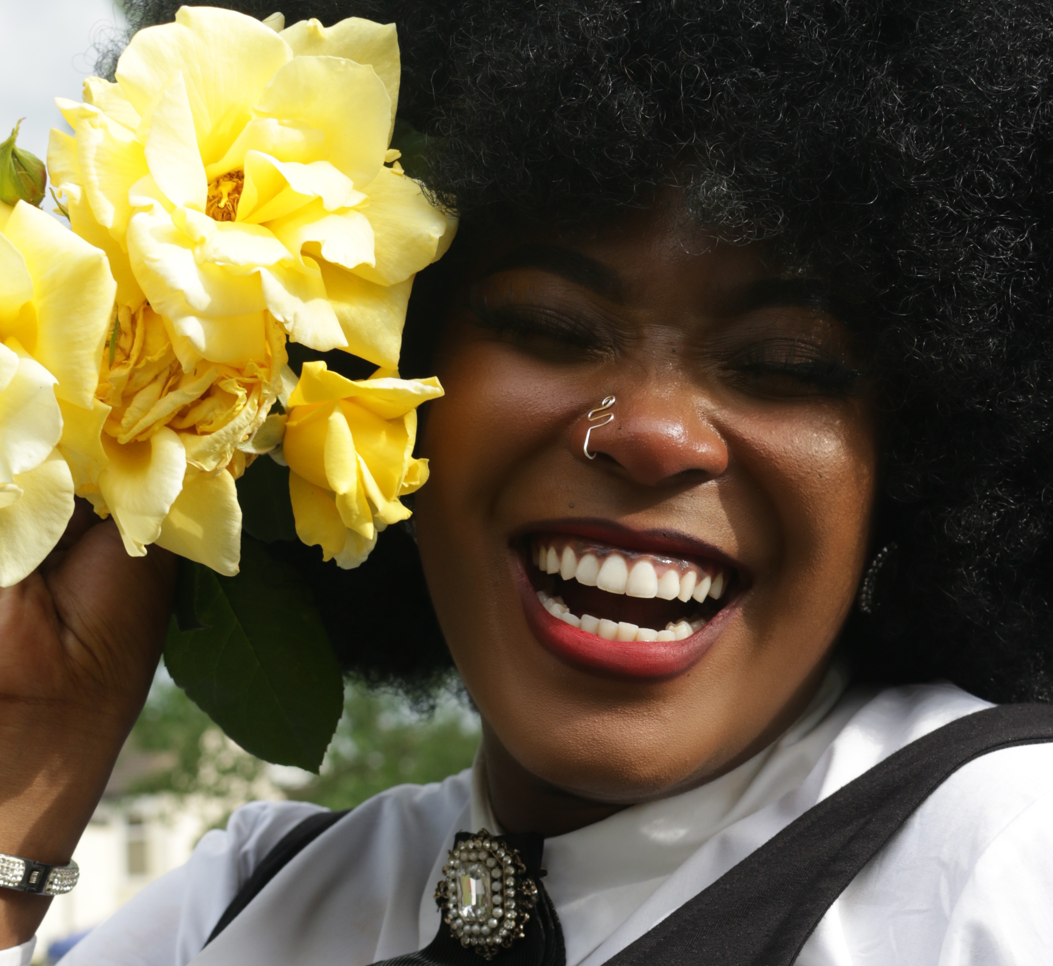 Tight shot of a Black woman with an afro smiling big while holding big yellow flowers next to her face.