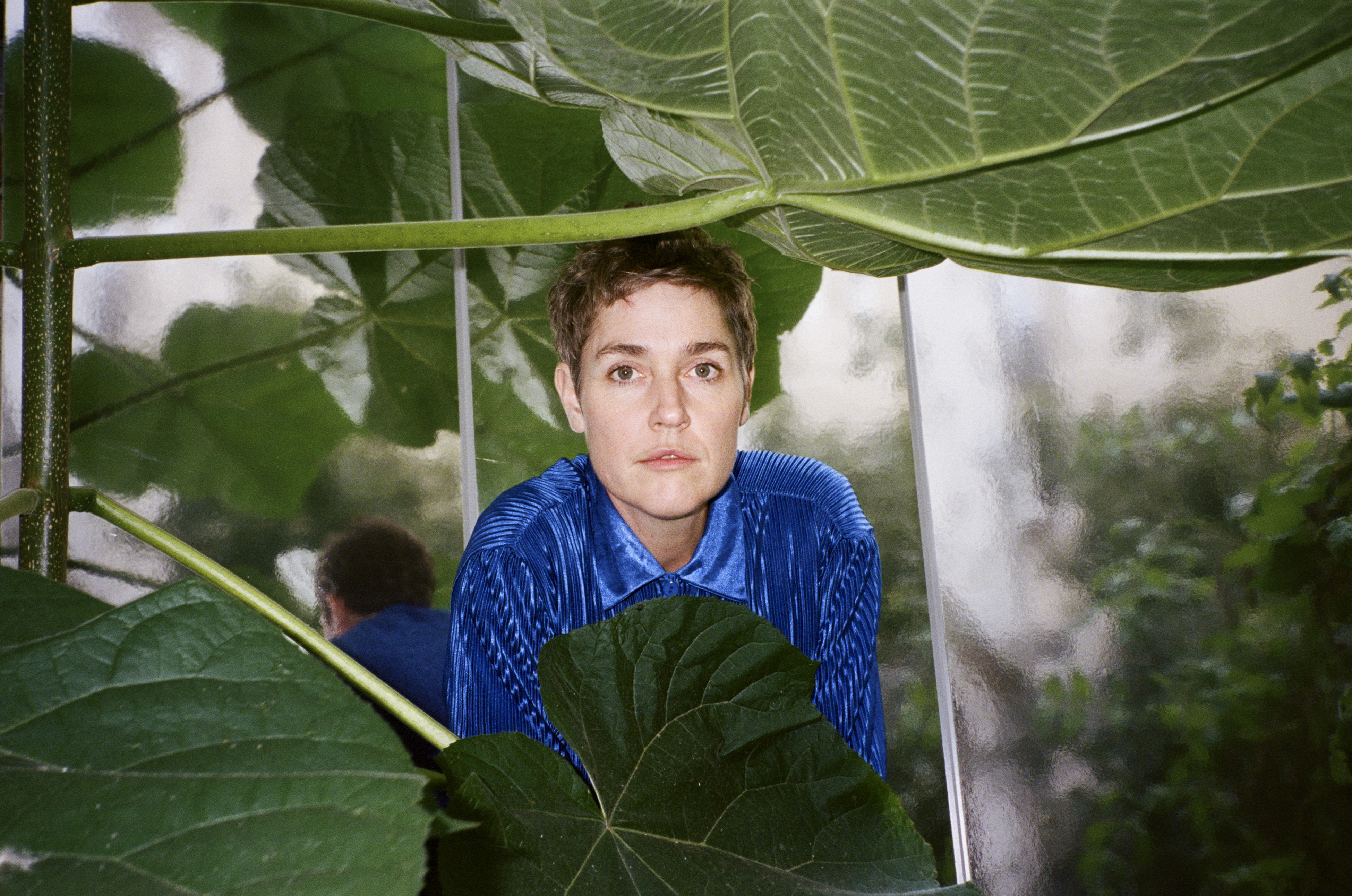 Portrait of Laura Steiner, songwriter and multi-instrumentalist of storm lily, in a blue velvet shirt in the background with leaves from a wide-leaf plant in the foreground.