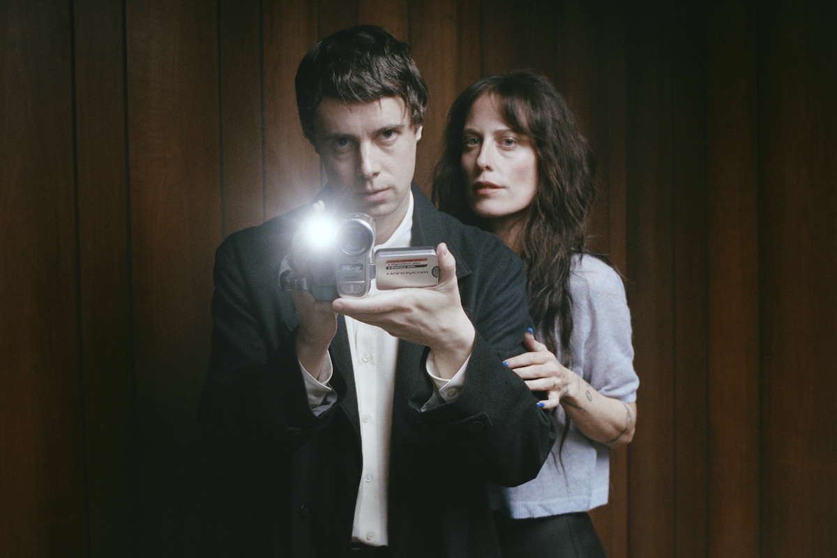The duo of Boy Harsher taking a mirror photo. A man in a black suit jacket, taking a photo in a mirror with a brunette woman standing behind him, holding his arm. Minimal with wood paneling behind them.