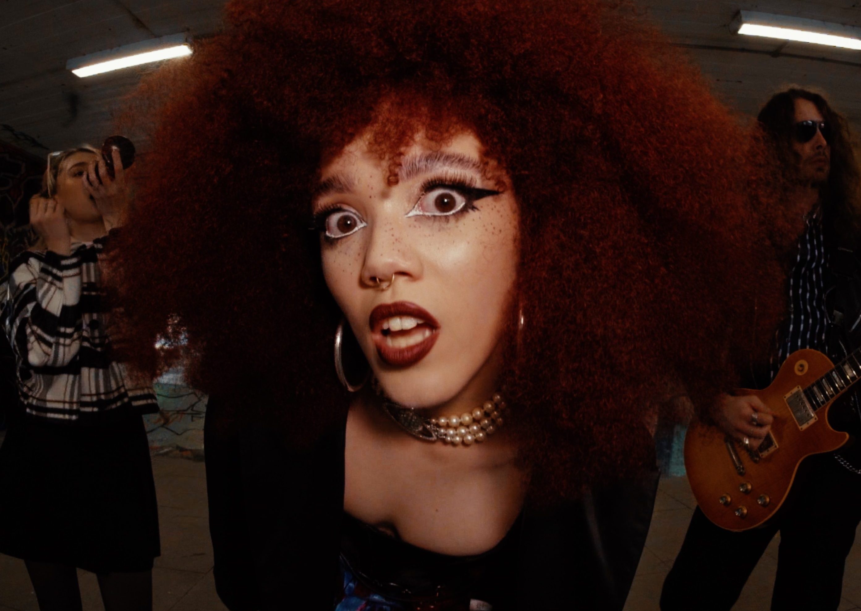 Music artist Tiny staring wide into the camera at close range. She has a dark red afro with two other musicians behind her.
