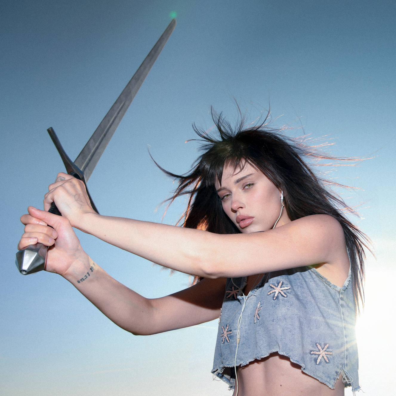Woman with long dark hair and bangs wields a long silver sword while in denim crop top