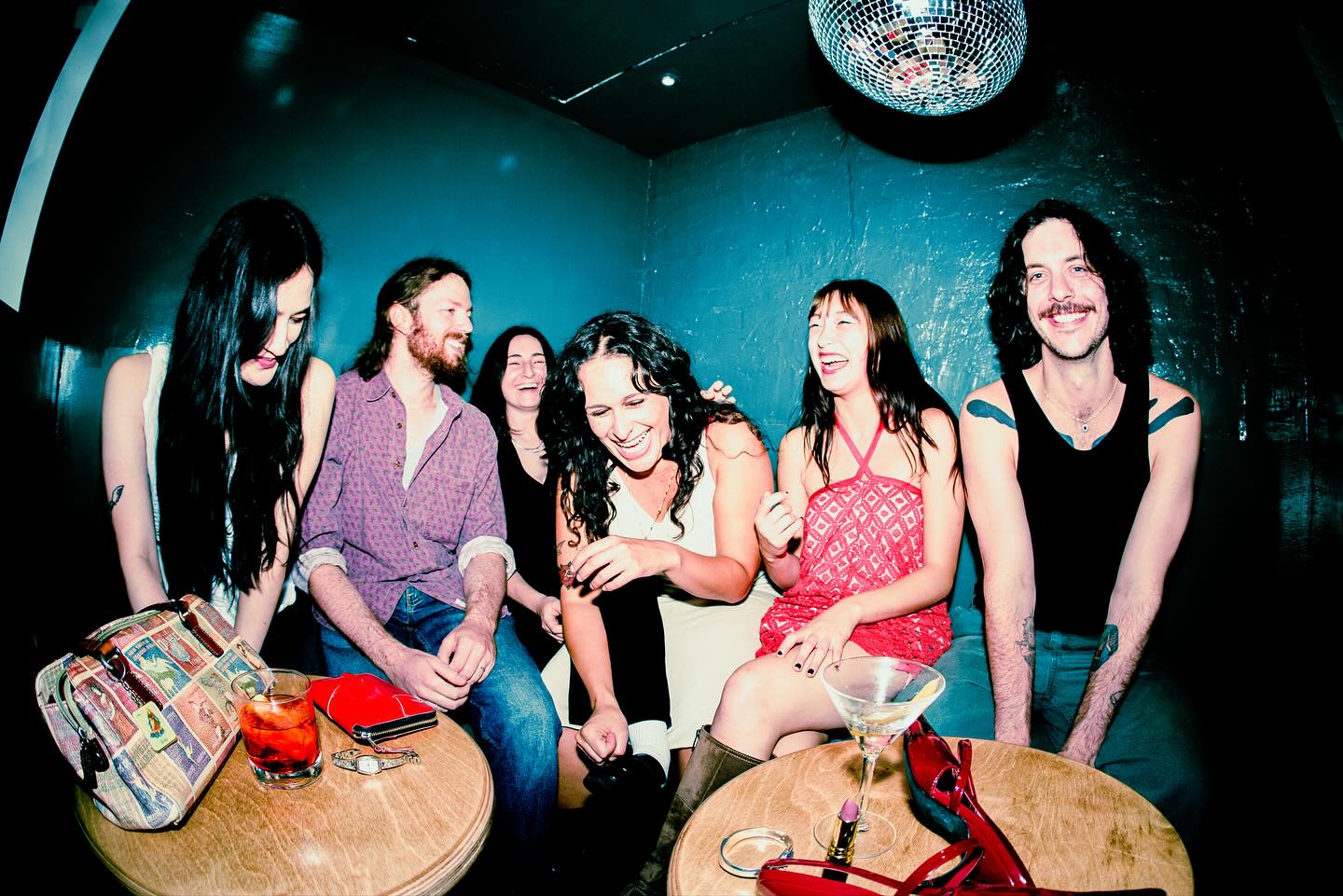 Six piece band partygirl sitting in a booth in a dark lounge, laughing with a disco ball above their heads. Purse, heels, and a martini glass on the table.