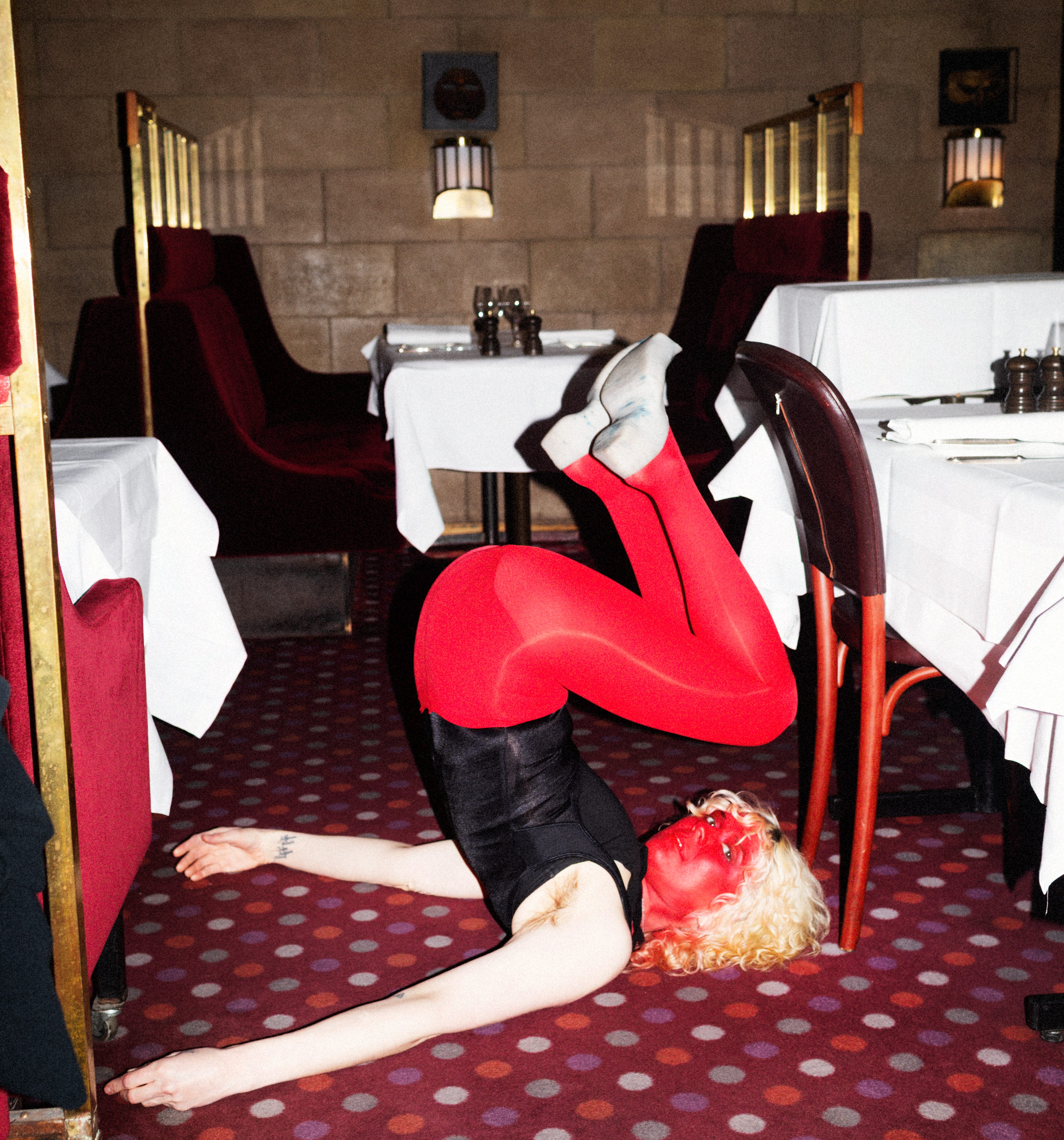 Woman with red painted face and short blonde curly hair is upside down on a maroon hotel carpet in red tights and a black unitard.