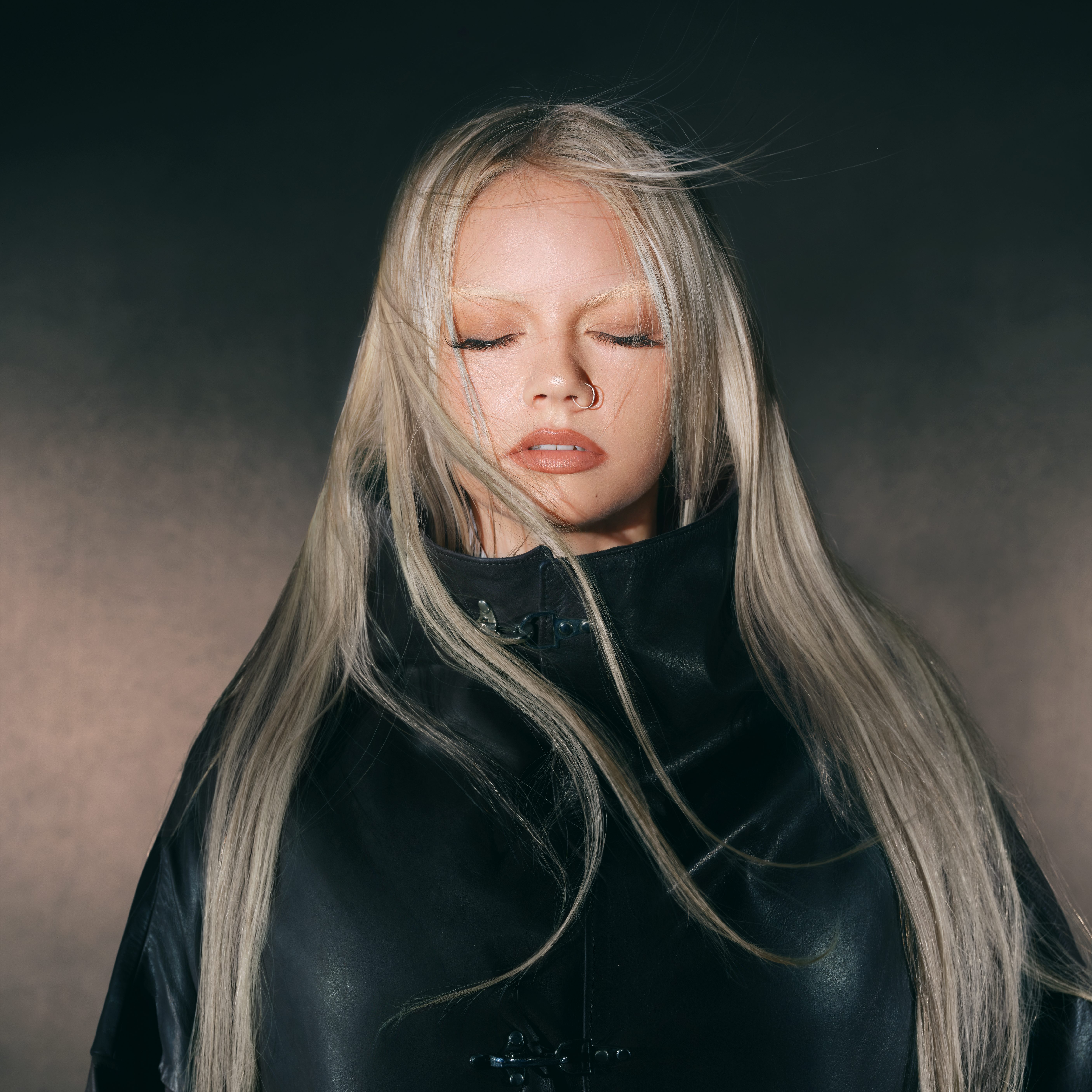 Decorative portrait of singer CHINCHILLA with long platinum blonde in a high collar black leather jacker while her eyes are closed.